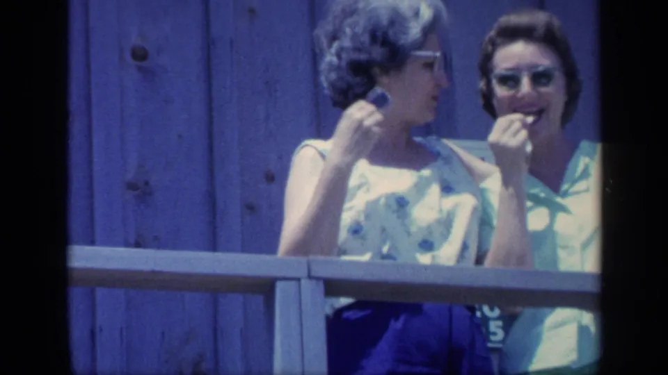 1968 Florida home-movie frame of two women behind a wooden railing, one in glasses and a sleeveless patterned top as they eat snacks.