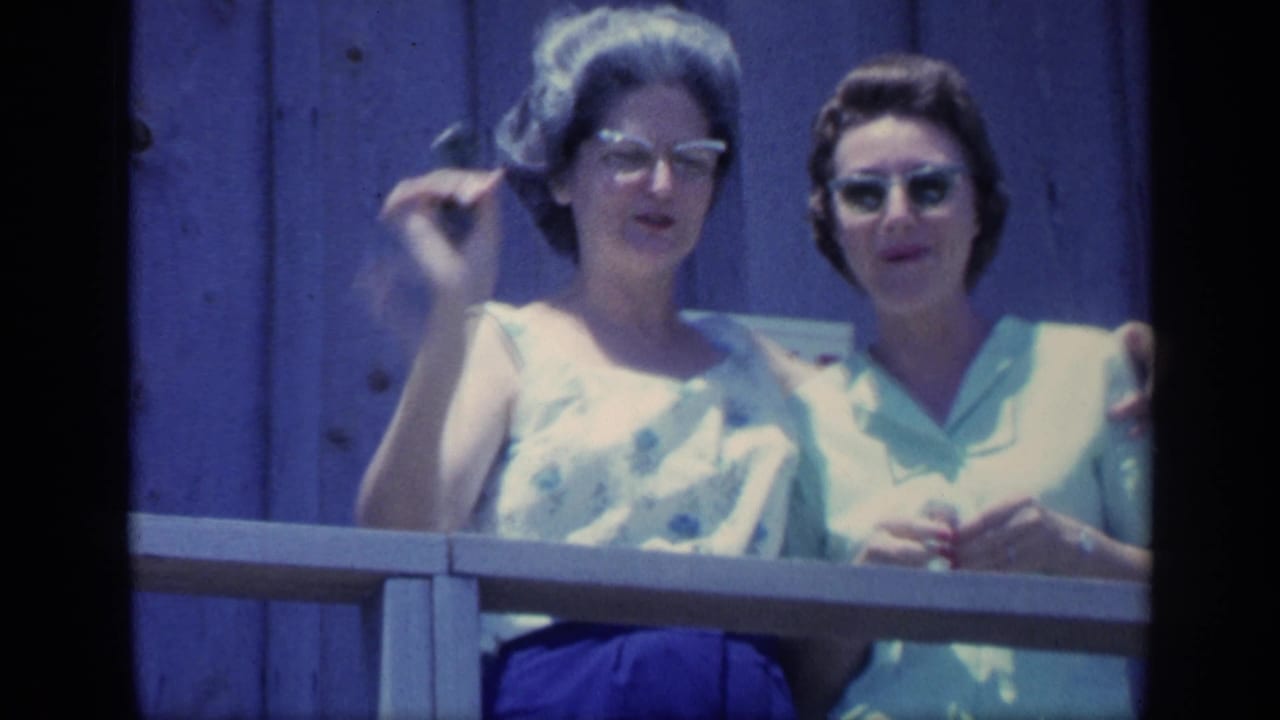 Two women smile behind a railing, with one raising a small snack or utensil.