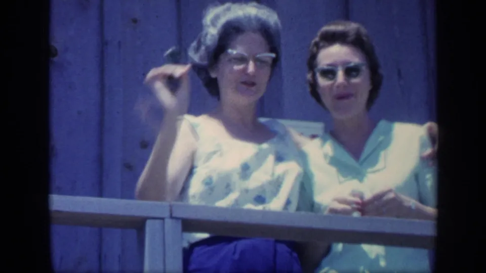 1968 Florida home-movie frame showing two women smiling at a wooden deck railing, one wearing glasses and the other wearing sunglasses.
