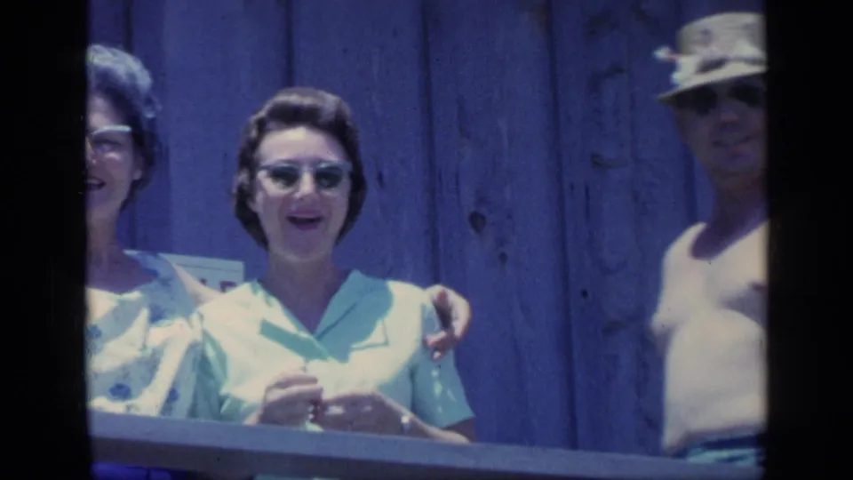 1968 Florida home-movie frame of a woman in sunglasses at a wooden railing as a shirtless man in a straw hat joins from the side.