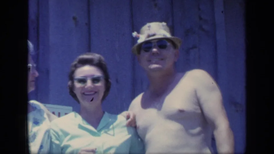 1968 Florida home-movie frame of a shirtless man in sunglasses and straw hat standing beside a smiling woman at a wooden deck railing.