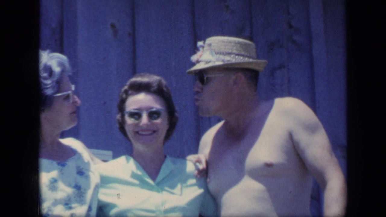 1968 Florida home-movie frame of two women and a shirtless man in a straw hat smiling on a wooden porch.