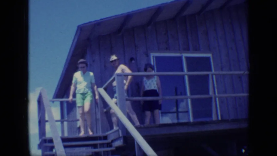 1968 Florida home-movie frame of adults on a raised wooden porch, with one person starting down the stairs under blue sky.