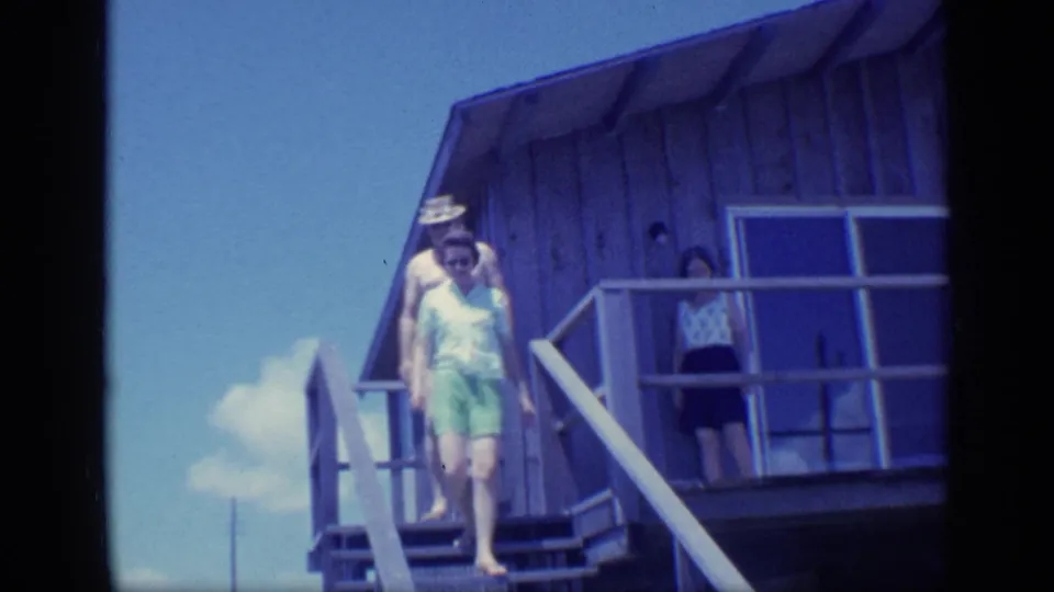 1968 Florida frame showing an adult descending outdoor stairs while others stand near the railing of a wooden beach house.