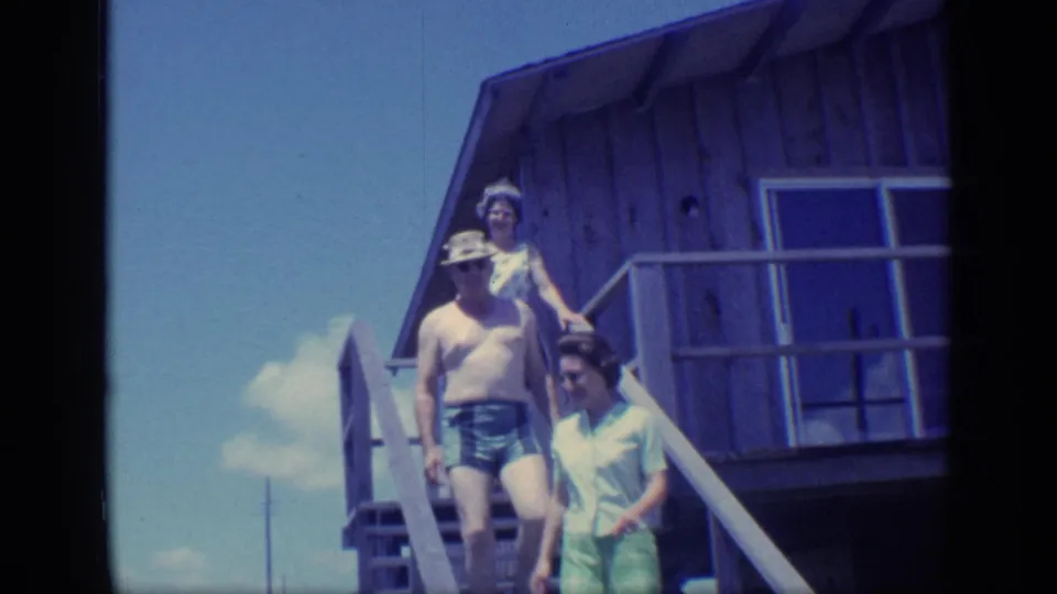 The group moves down the deck stairs, with a shirtless man in swim trunks leading and two women following in bright daylight.