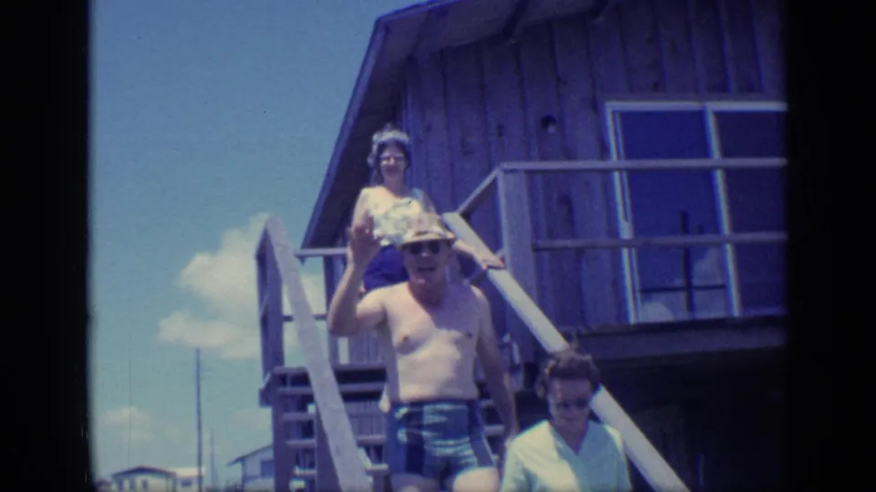 1968 Florida home-movie frame of adults on wooden beach-house stairs, with a shirtless man waving toward the camera.