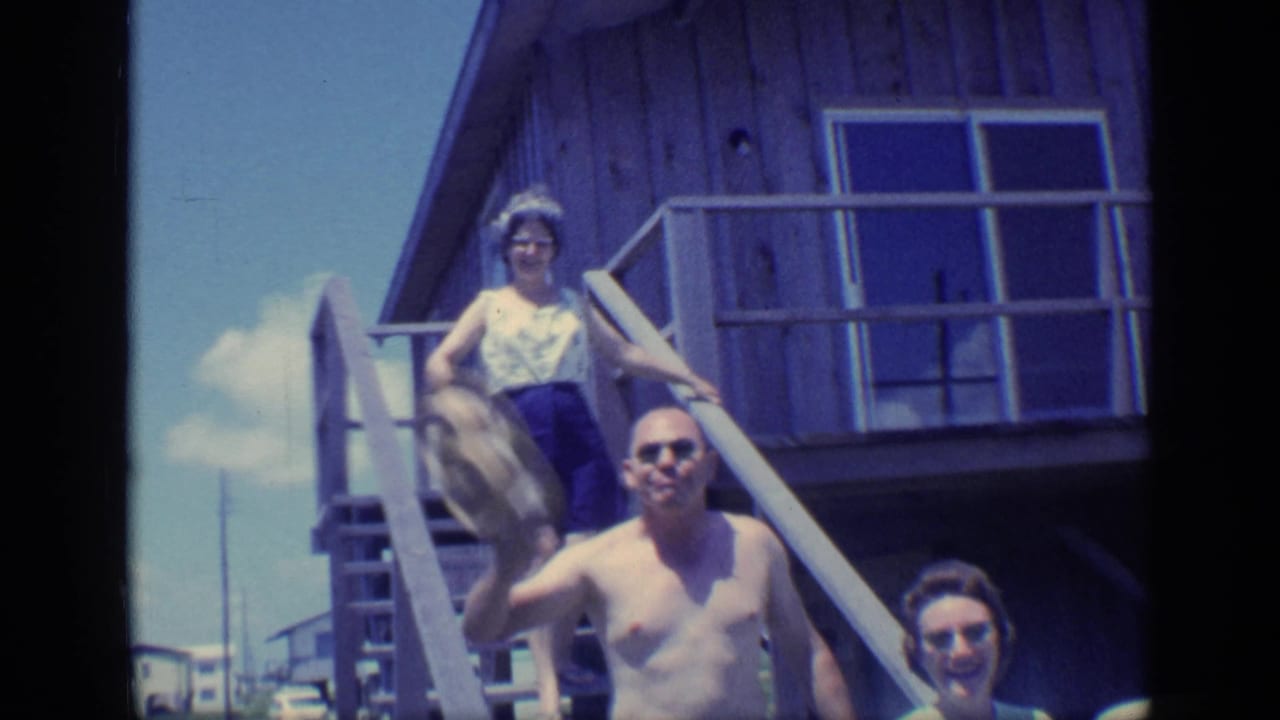 A shirtless man waves while women stand and descend on beach-house stairs.