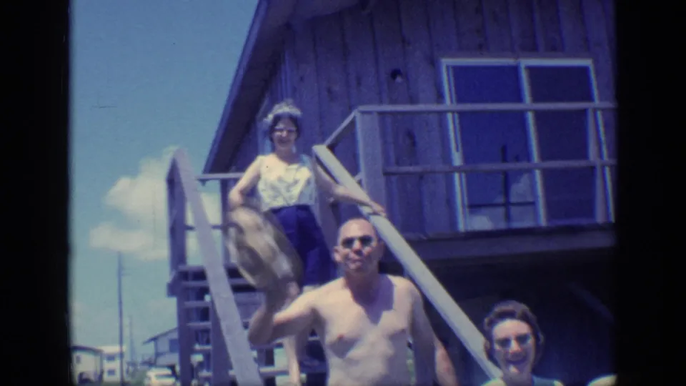 1968 Florida beach-house scene with a shirtless man in sunglasses waving and women on the stairs behind him.
