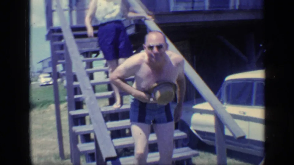 1968 Florida home-movie frame showing a man descending beach-house stairs, with a woman above and a light-colored car nearby.