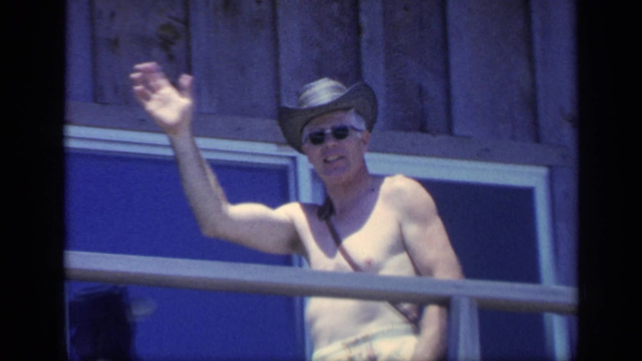Shirtless man in sunglasses and cowboy hat waves from a wooden porch.