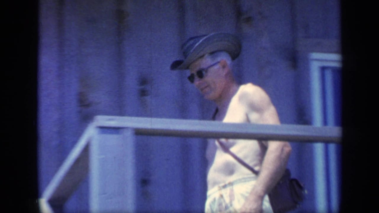 Man in hat and sunglasses stands beside a porch railing near wood siding.