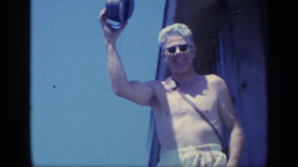 The man smiles toward the camera while lifting his hat, with blue sky and the edge of a wooden building behind him.