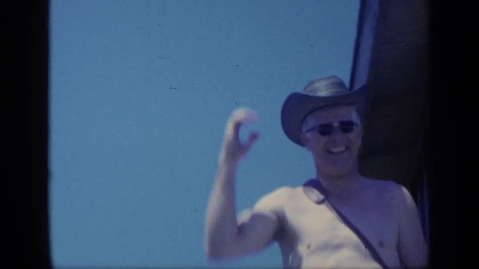The man smiles and waves again in his cowboy-style hat and sunglasses, framed by blue sky and a dark doorway edge.