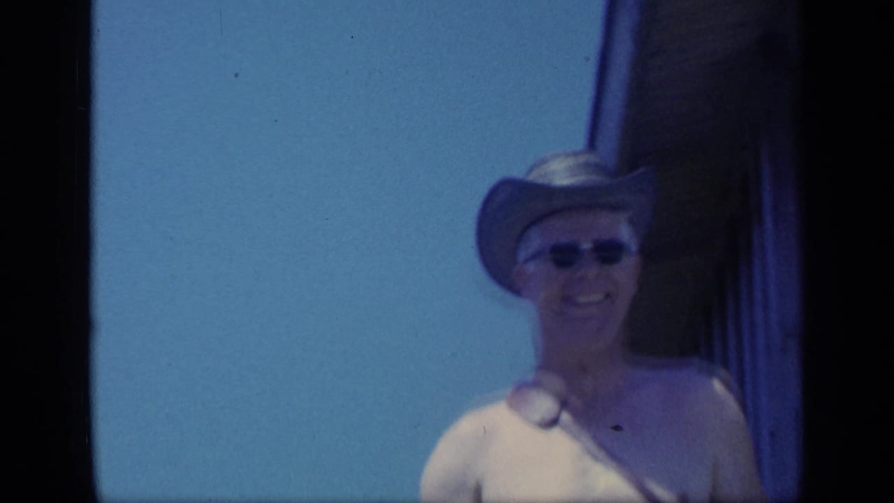Shirtless man in a cowboy-style hat and sunglasses stands near wooden stairs.
