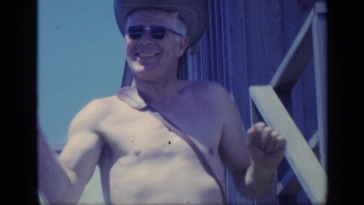 Shirtless man in sunglasses smiles while holding a hand near porch steps.