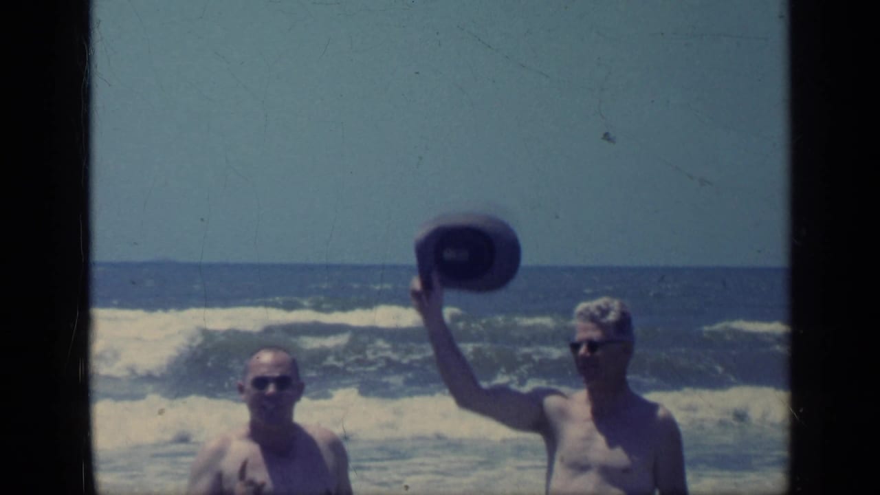Two men in sunglasses stand at the beach as one raises a hat near the surf.