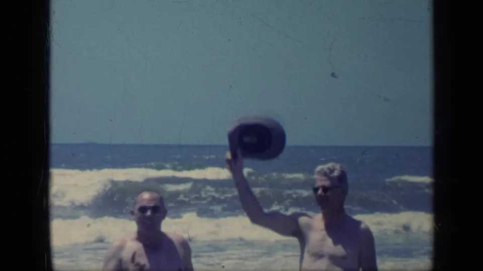 1968 Florida home-movie frame of two adult men at the shoreline, one lifting a hat while waves break behind them.