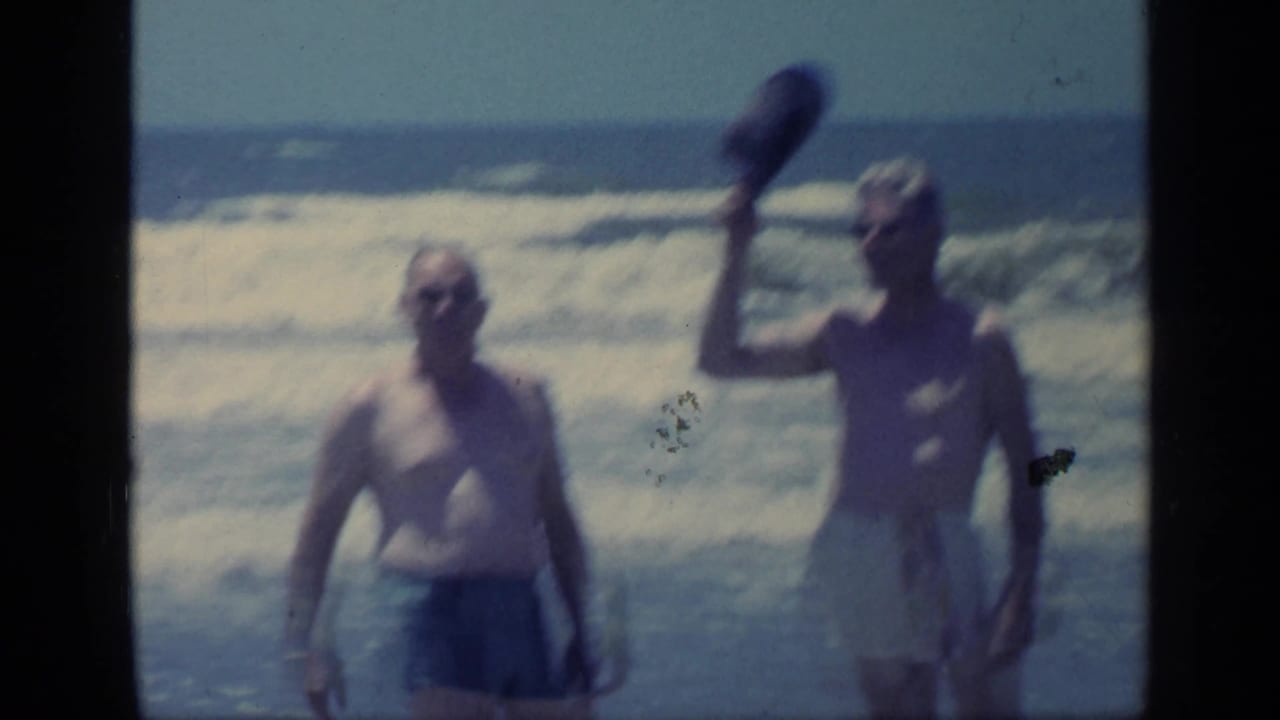 Two adult men in swim trunks stand before breaking waves in Florida, with one man raising a hat toward the camera.