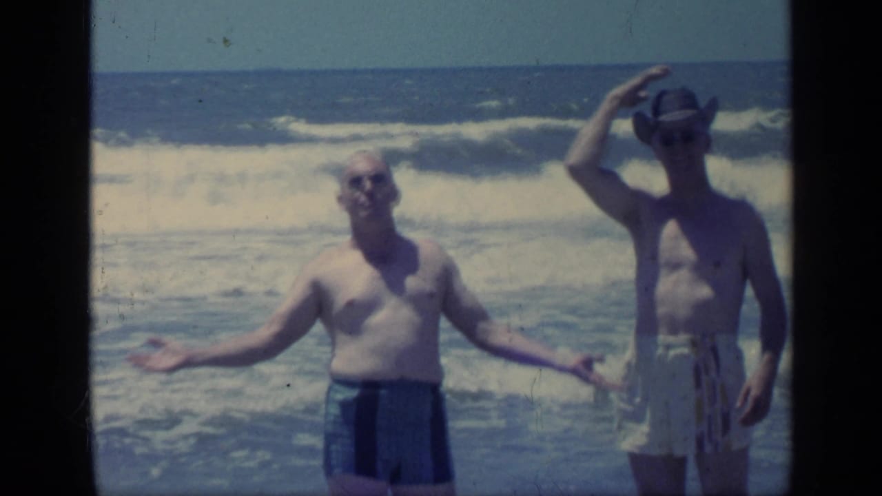 In a 1968 Florida beach frame, one man opens his arms toward the camera while another adjusts or holds a hat above his head.