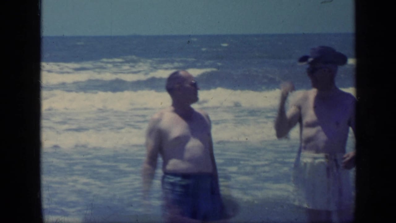 Two adult men in swimwear stand at a Florida shoreline in 1968 as one turns and gestures while surf rolls behind them.