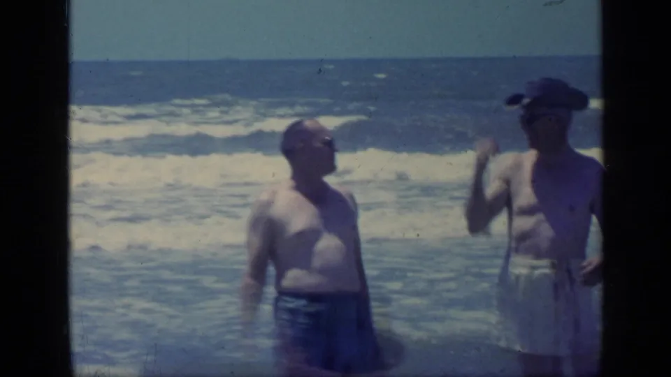 Two adult men in swimwear stand at a Florida shoreline in 1968 as one turns and gestures while surf rolls behind them.