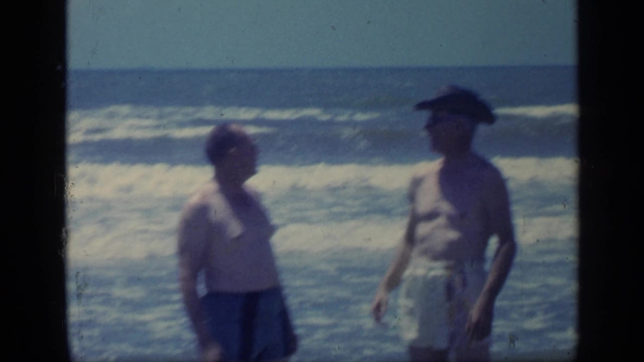 In supported 1968 Florida home-movie footage, two adult men in swimwear stand by rolling ocean waves, one wearing a dark hat.