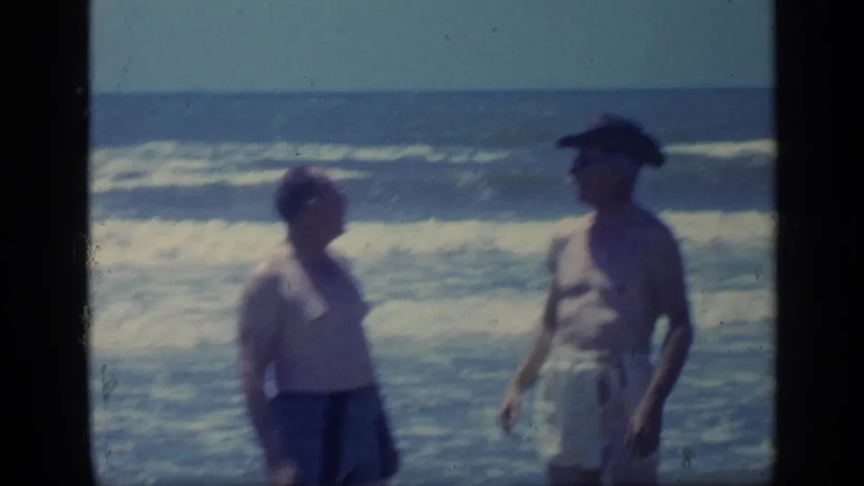 In supported 1968 Florida home-movie footage, two adult men in swimwear stand by rolling ocean waves, one wearing a dark hat.