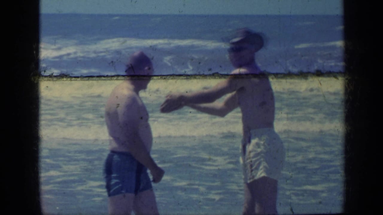 Two adult men stand beside the ocean in the Florida home-movie scene, with one man in light trunks and a hat gesturing near the surf.