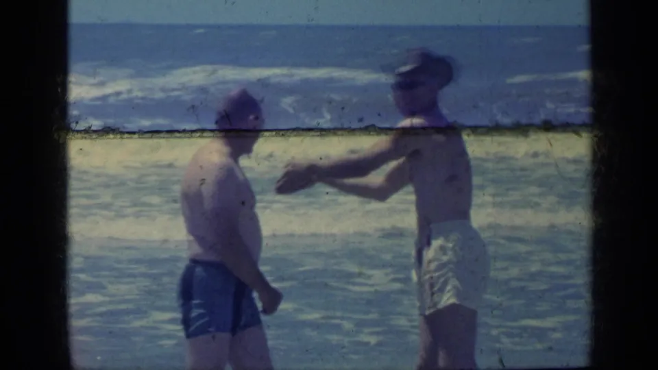 Two adult men stand beside the ocean in the Florida home-movie scene, with one man in light trunks and a hat gesturing near the surf.