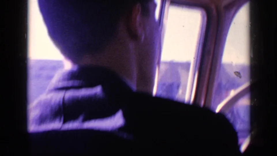 A man in a dark jacket is seen from behind driving a car in bright daylight, filmed from inside the vehicle on 8mm.