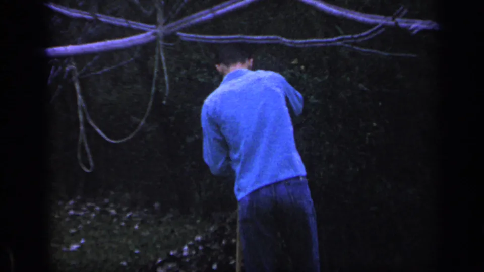 A man in a blue shirt stands with his back to the camera in a dark outdoor area, near visible hanging lines or branches.