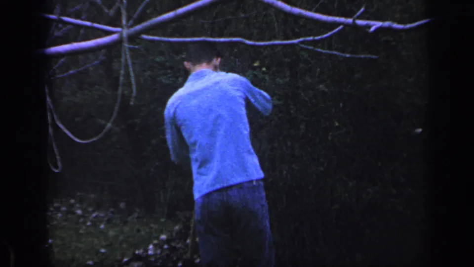 In this 1968 Florida 8mm frame, a man in a blue long-sleeved shirt stands with his back to the camera in a dim wooded backyard.