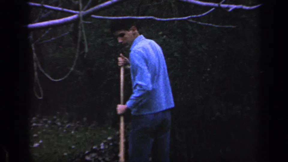 A 1968 Florida home-movie frame shows the man holding a rake upright in a shaded backyard with leaves and foliage around him.