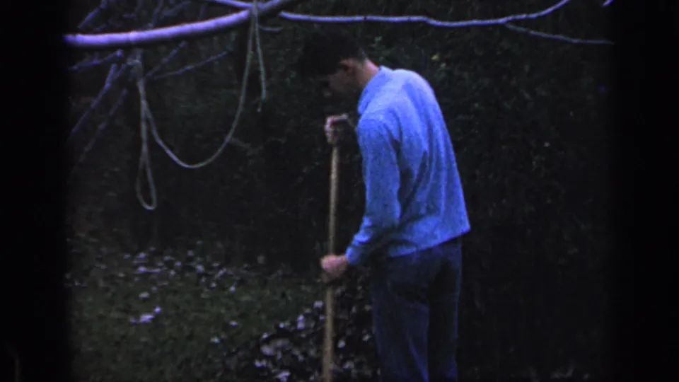 The man leans over the rake in the wooded Florida yard, framed by dense foliage, scattered leaves, and a looping rope above.