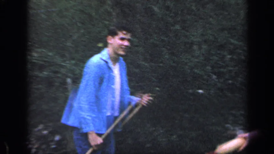 1968 Florida home-movie frame of a smiling man in a blue shirt holding a long-handled yard tool in dense greenery.