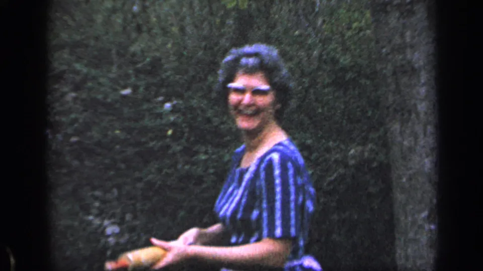 1968 Florida home-movie frame showing a smiling woman in sunglasses and a striped dress holding a hand yard tool near greenery.