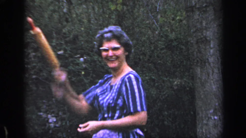 Adult woman in a blue-and-white striped dress smiles and waves toward the camera in 1968 Florida home-movie footage.