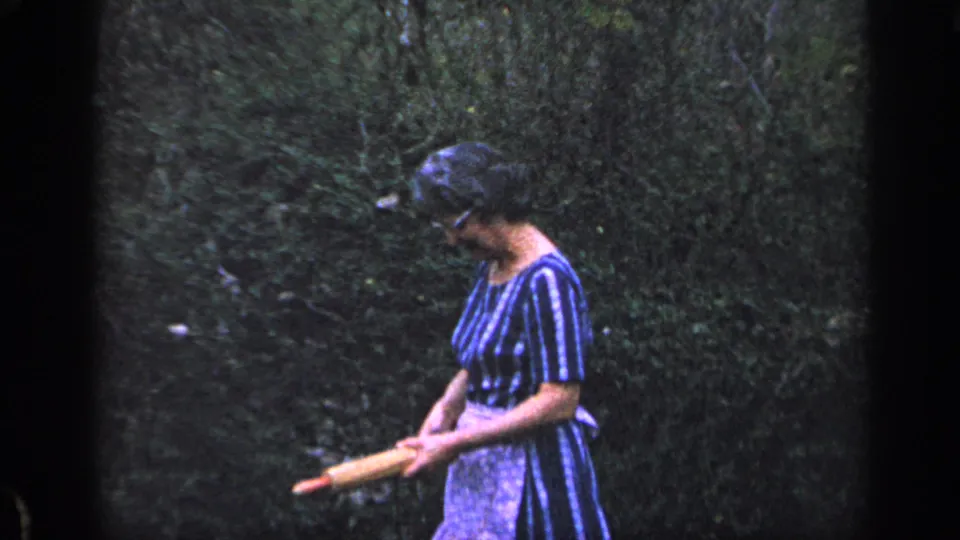 The woman stands in profile in the garden, looking down while holding a loaf of bread in 1968 Florida home-movie footage.