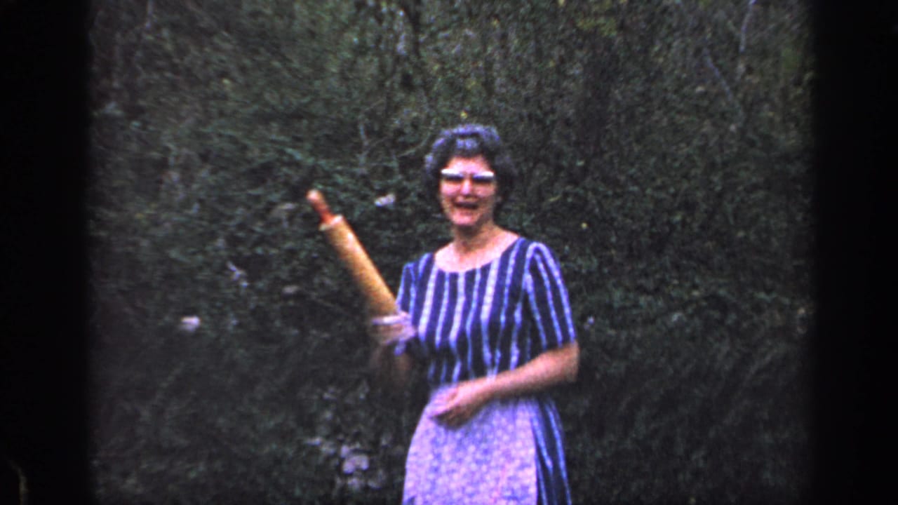 Woman in glasses stands in a leafy yard holding up a rolling pin.