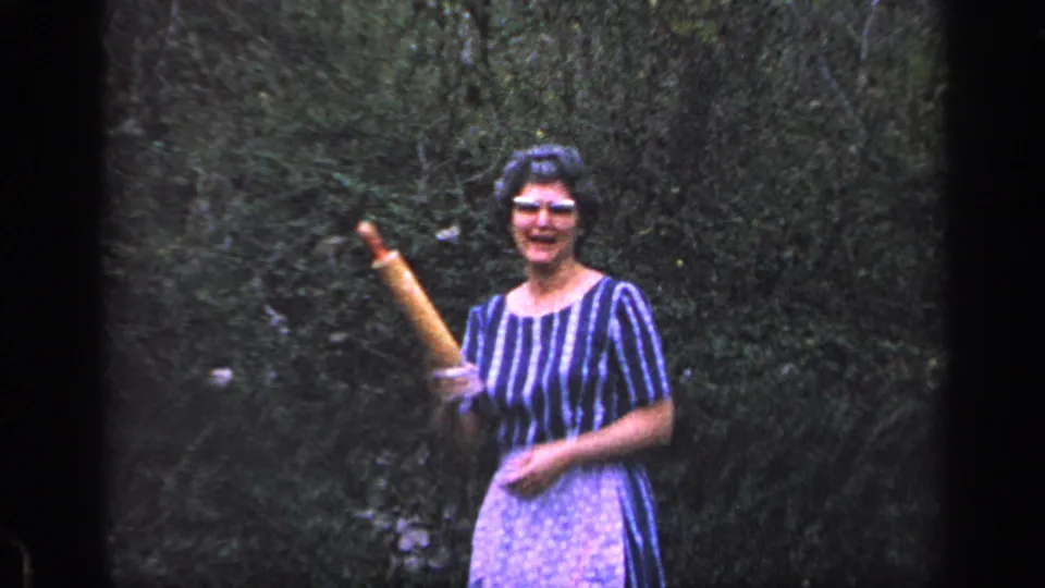 In 1968 Florida home-movie footage, a woman in glasses, striped dress and apron smiles while raising a wooden rolling pin in a yard.