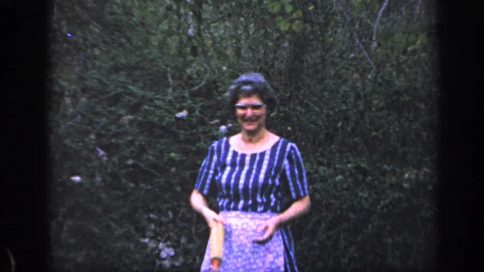 The woman stands before dense greenery in Florida, 1968, smiling at the camera with a wooden rolling pin held against her apron.