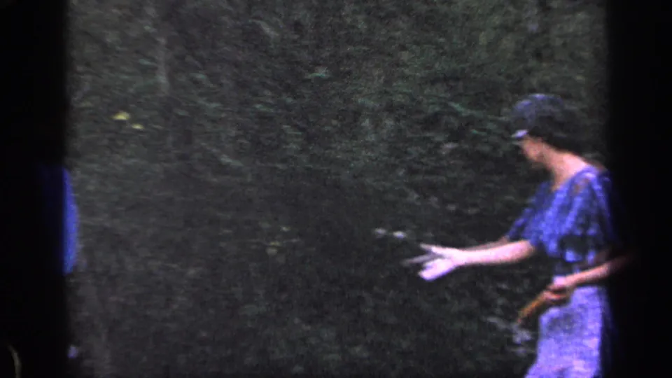The woman turns partly away in the leafy yard, extending one hand while holding the rolling pin in the other during the 1968 home-movie moment.
