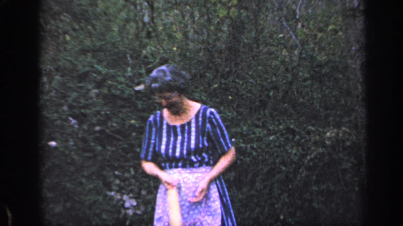 Older woman in a striped dress and apron looks down while holding a rolling pin in a wooded yard.