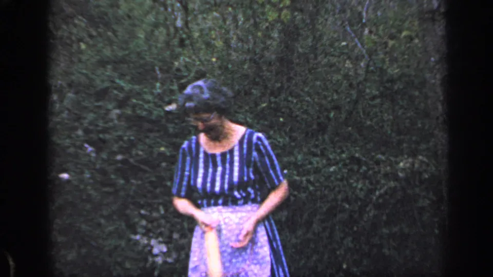 In 1968 Florida home-movie footage, an older woman in a striped dress and apron looks down while holding a rolling pin outdoors.