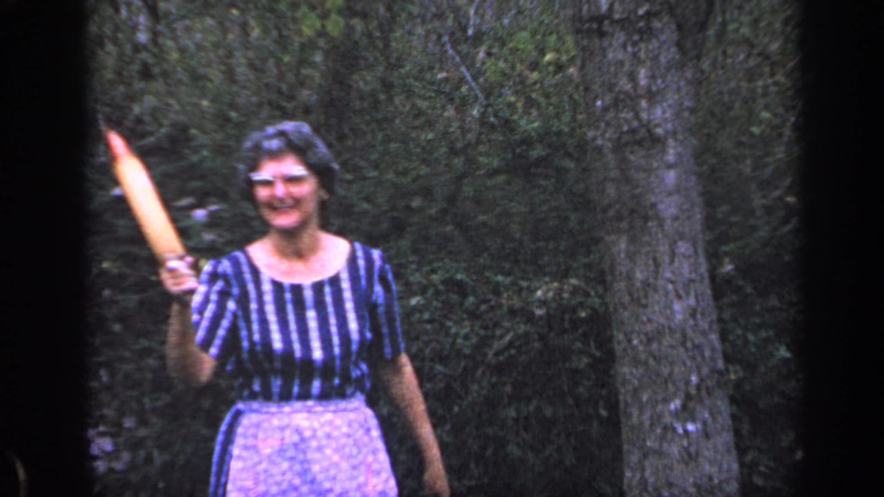 Smiling older woman raises a rolling pin beside a tree trunk in a wooded outdoor setting.