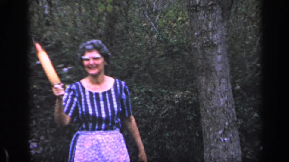 The woman smiles toward the camera and raises a rolling pin in a wooded Florida yard, consistent with the 1968 home-movie metadata.