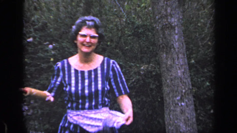 An older woman in a striped dress and pale apron smiles toward the camera in a wooded Florida yard in this 1968 home-movie frame.