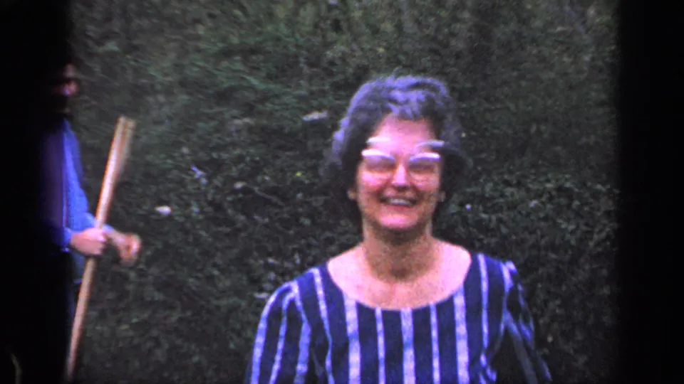 Close 1968 Florida home-movie frame of the smiling woman in eyeglasses, with a partly visible person holding the rolling pin nearby.