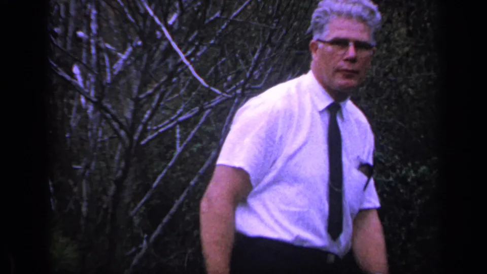 A man in a white short-sleeved shirt and dark tie stands in a Florida yard in this 1968 home-movie frame.