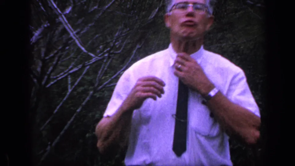 The man adjusts his dark tie in a 1968 Florida home-movie frame, with branches and greenery behind him.